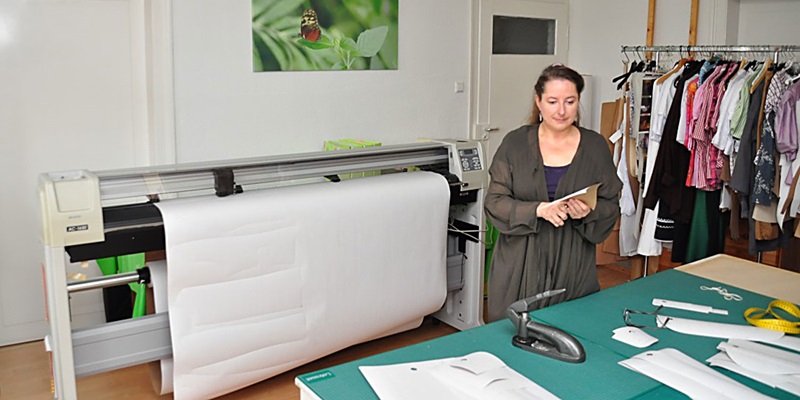 Karin Widmann in ihrem Atelier am Zuschneidetisch, im Hintergrund Rollenplotter für Pappschablonen